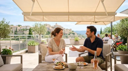 Un couple vu de dos profite d'une terrasse moderne sous un store banne déployé, l'un assis avec une tablette, l'autre debout regardant le jardin, lumière tamisée par la toile créant une atmosphère apaisée malgré le soleil éclatant à l'extérieur