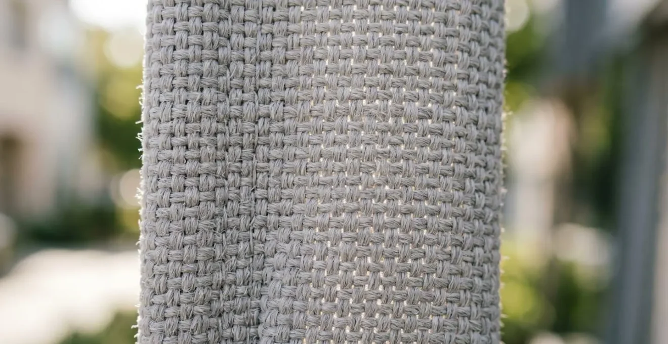 Gros plan extrême sur la texture tressée d'une toile de store acrylique, avec trame visible et grammage épais, lumière naturelle filtrant à travers les fibres créant un jeu d'ombre et de lumière